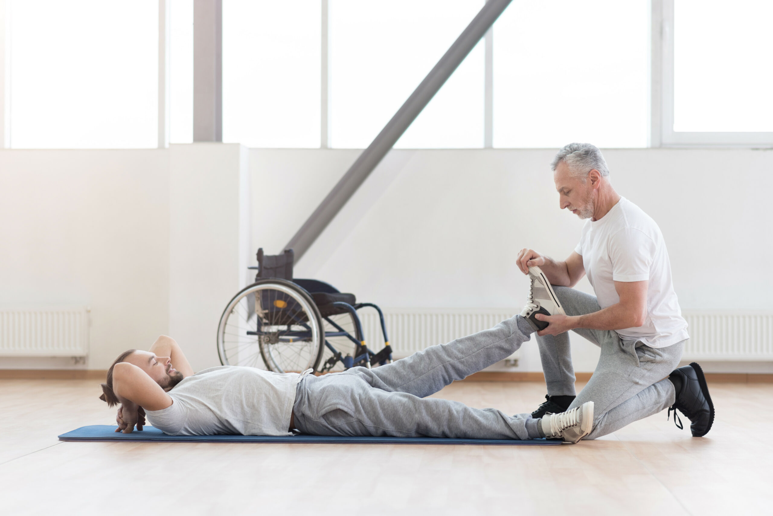 Doing my job properly. Confident skilled aged physical therapist helping the disabled man and providing a rehabilitation while expressing positivity and holding the leg of the patient